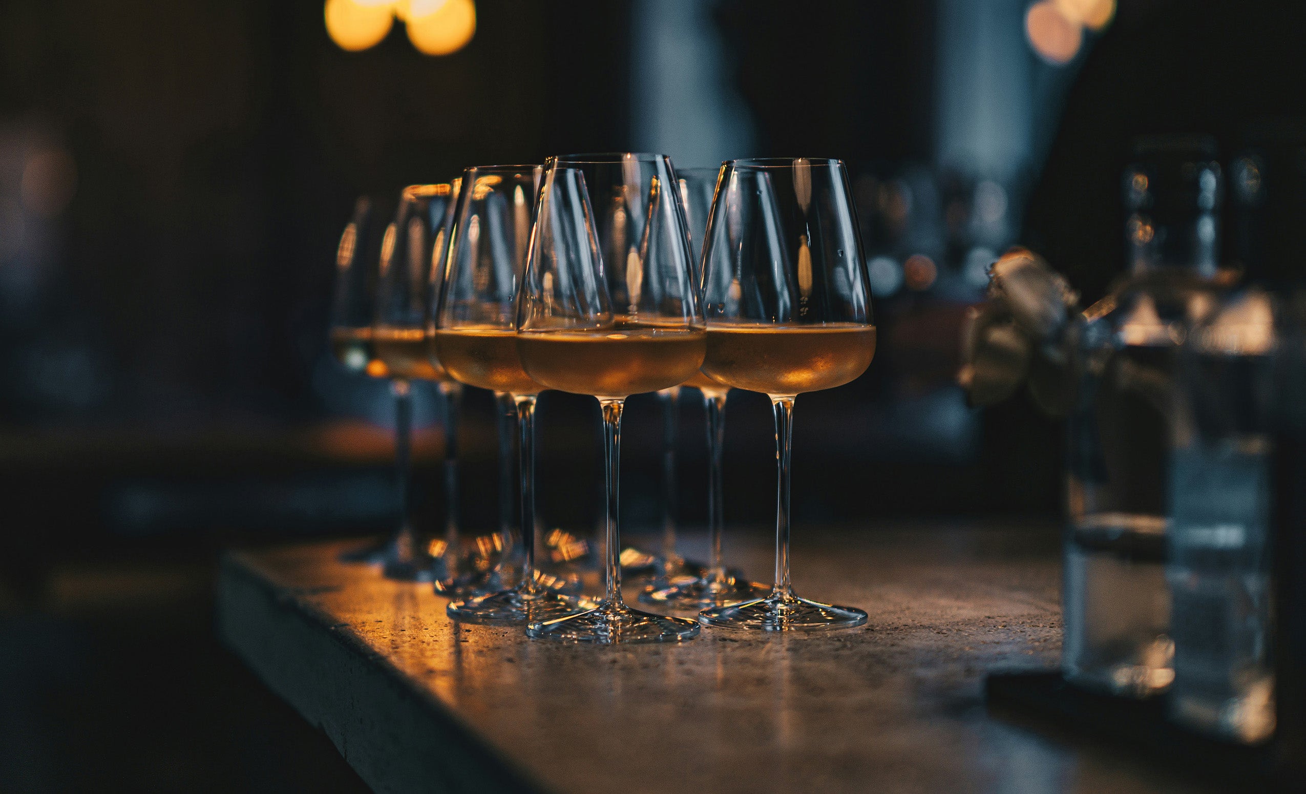 Row of wine glasses filled with pale gold wine on a dark bar counter, warm lighting, blurred bottle in background