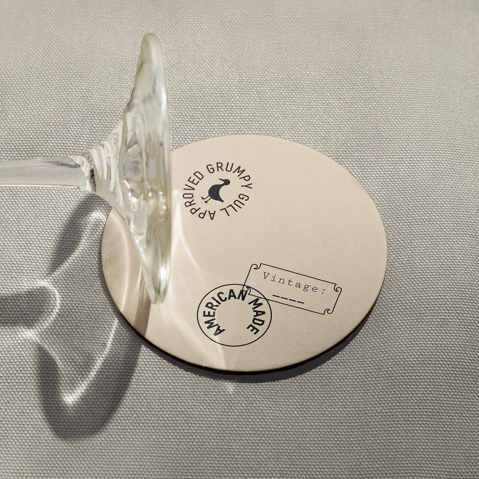 Round coaster stamped "Grumpy Gull approved, american-made," with wine glass stem resting on top