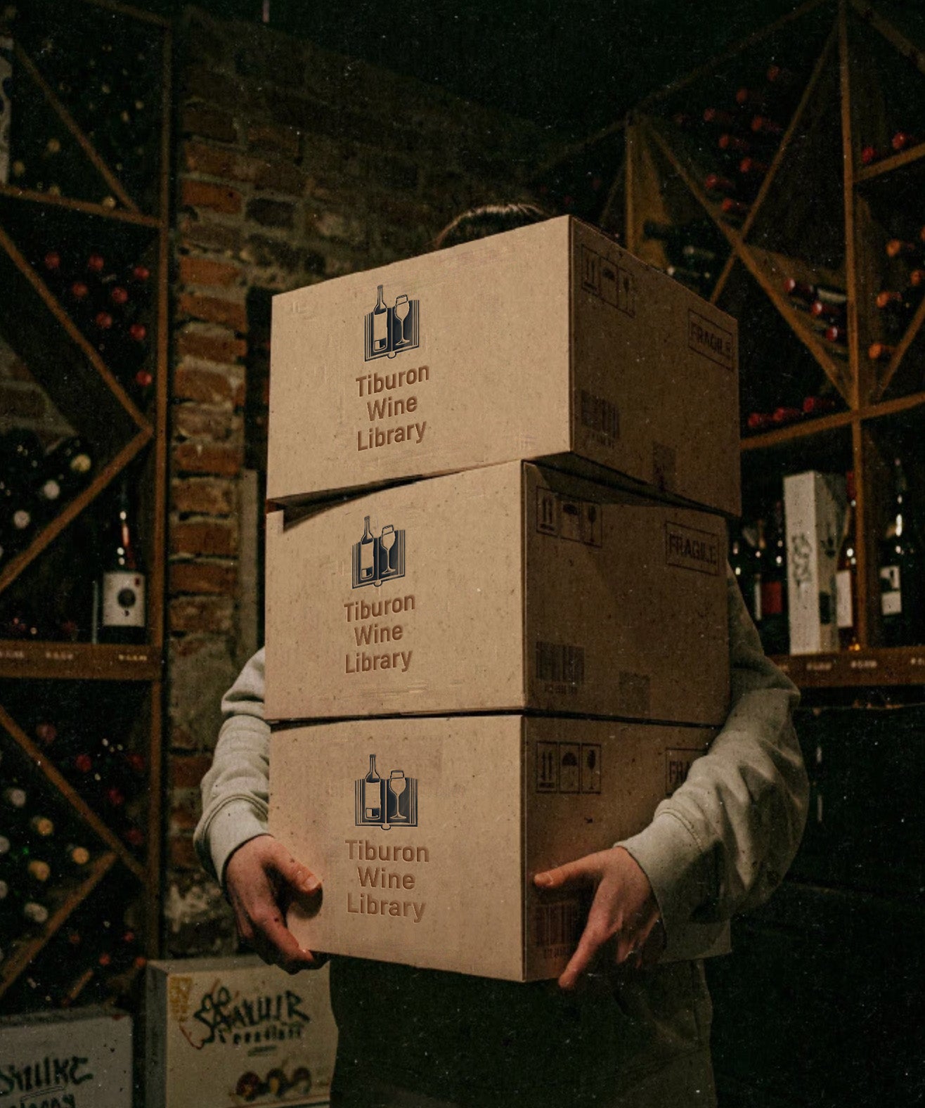 Person carrying three cardboard boxes labeled "Tiburon Wine Library" in a wine cellar